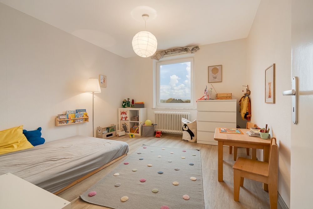 Helles Kinderzimmer mit Bett, Teppich, Tisch, Kommode und Spielzeugregal.