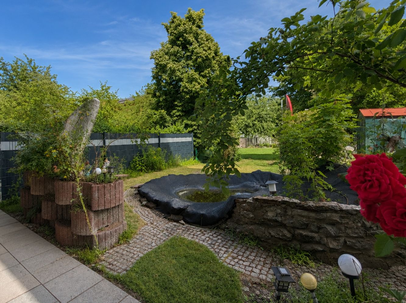 Garten mit kleinem Teich, Natursteinmauer, Pflasterweg und Blumen bei Sonnenschein.