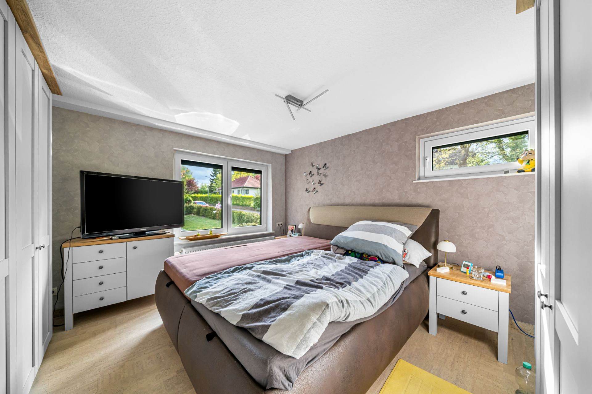 Helles Schlafzimmer mit Doppelbett, Fernseher, zwei Fenstern und Nachttischen mit Lampen.