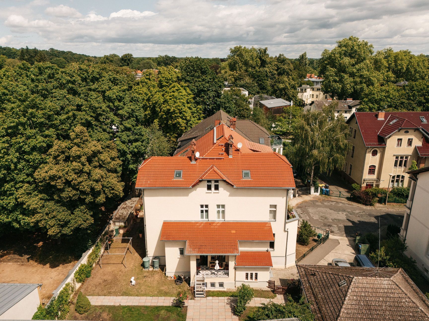 Großes Wohnhaus mit rotem Dach und kleinem Garten, umgeben von Bäumen und anderen Häusern.