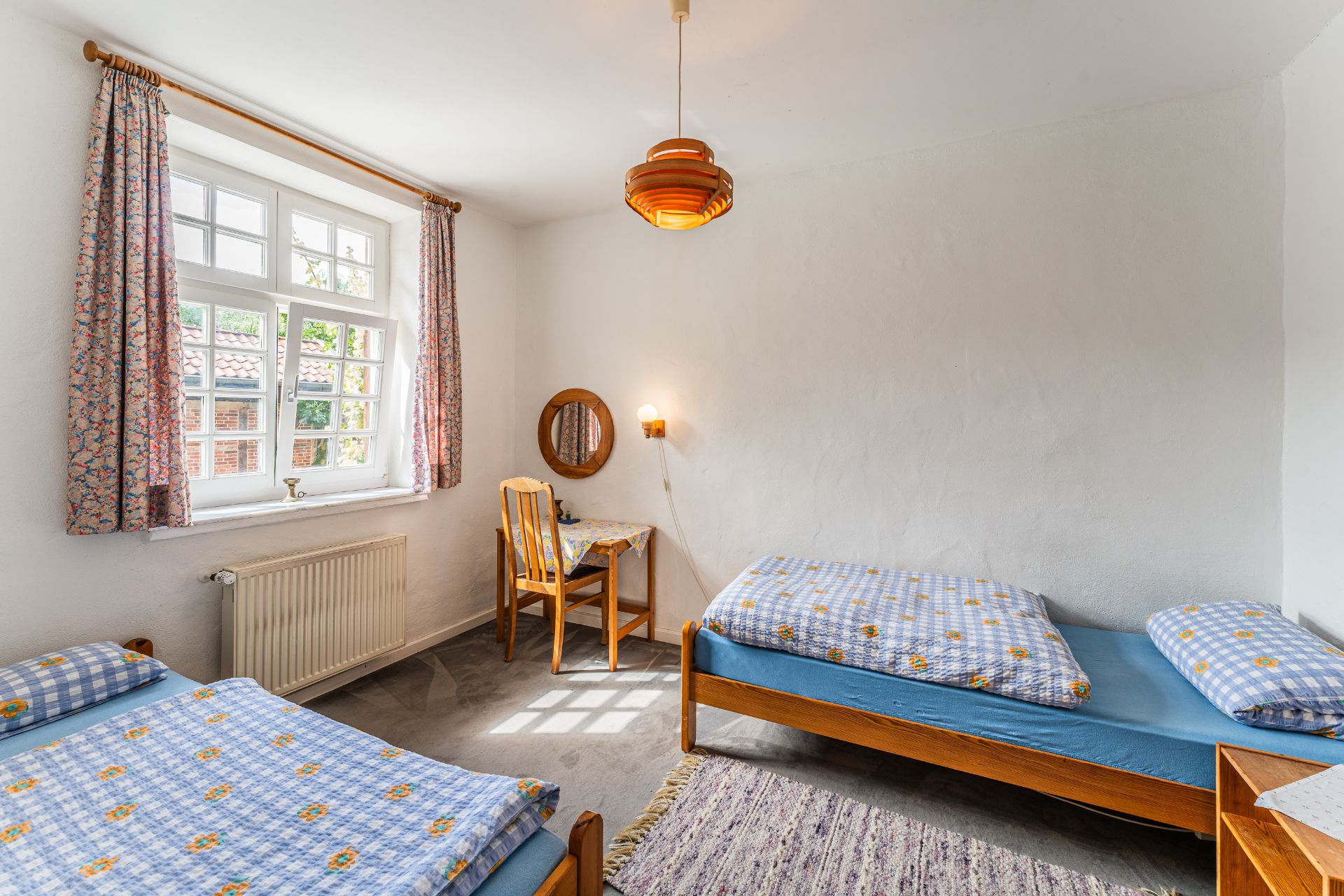 Helles Schlafzimmer mit zwei Einzelbetten, Schreibtisch am Fenster und blumengemusterten Vorhängen.