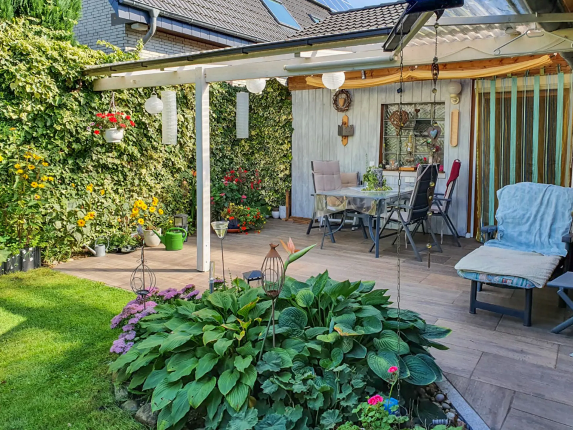Überdachte Terrasse mit Sitzgruppe, vielen Pflanzen und Gartenmöbeln im Garten eines Wohnhauses.