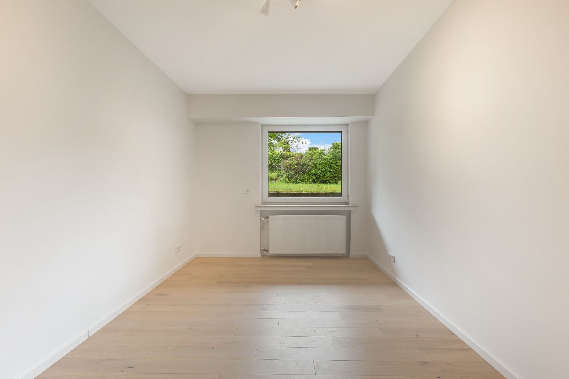 Helles, leeres Zimmer mit Holzfußboden und großem Fenster mit Blick ins Grüne.