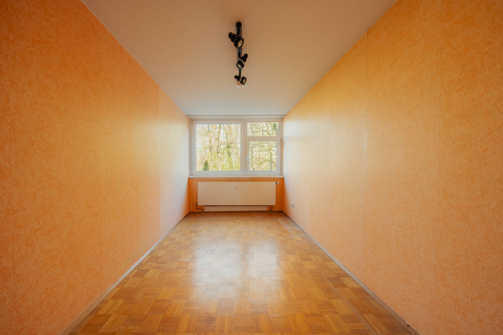 Langer, leerer Raum mit orangefarbenen Wänden, Parkettboden und großem Fenster am Ende.