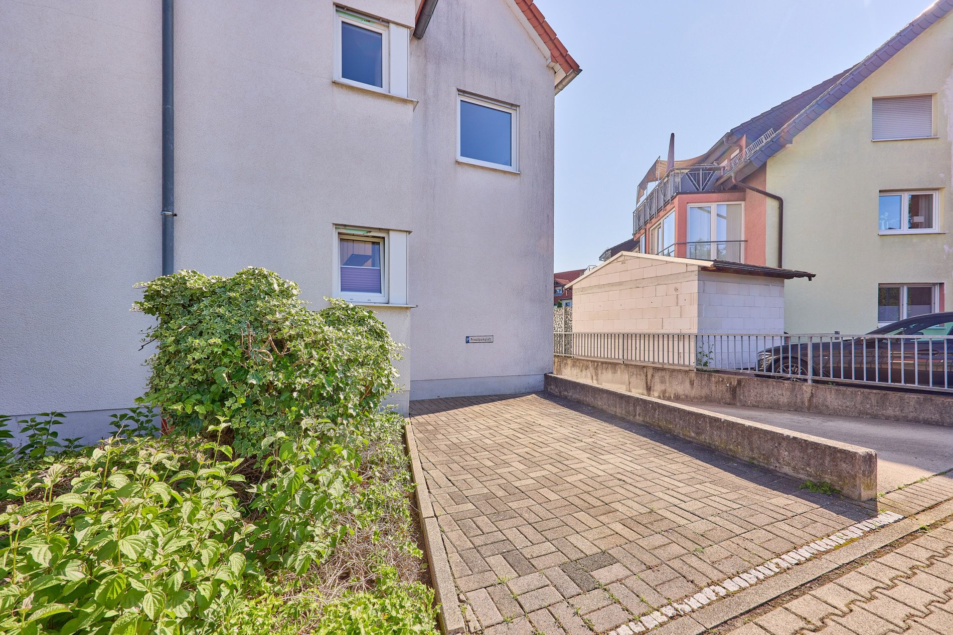 Gepflasterter Weg neben einem Wohnhaus mit Fenstern und Hecke im Vordergrund.