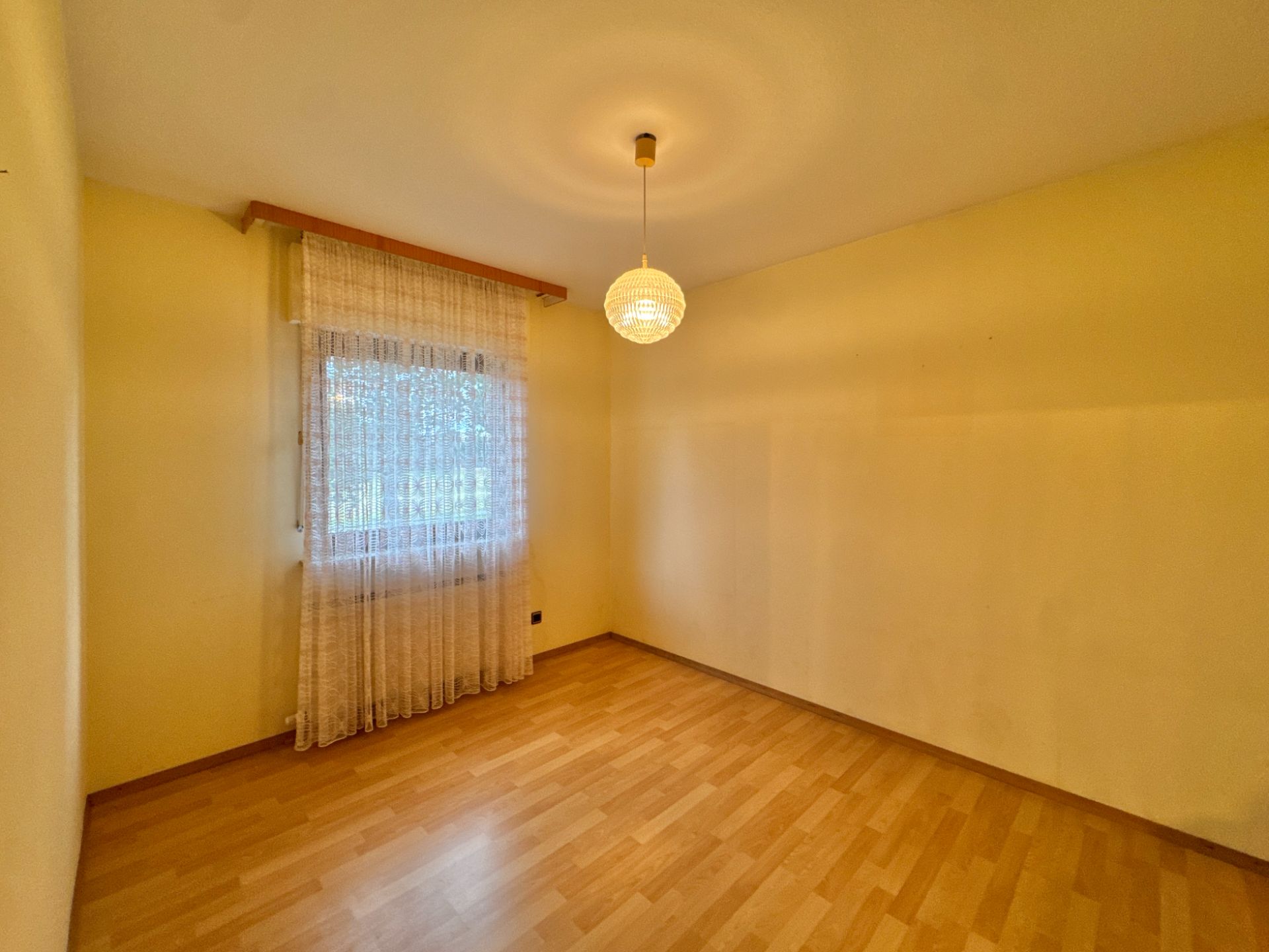 Leeres Zimmer mit Holzfußboden, gelben Wänden, Fenster mit Gardine und Deckenlampe.