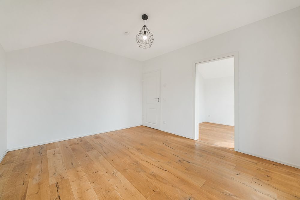 Leerer Raum mit weiß gestrichenen Wänden, Holzboden und moderner Deckenleuchte.