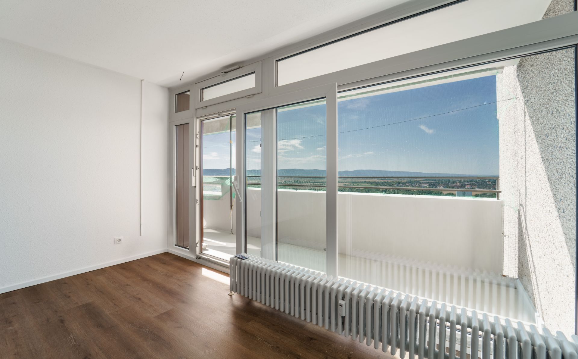 Heller Raum mit großem Fenster, Heizkörper und Zugang zu einem Balkon mit Ausblick ins Grüne.