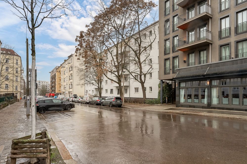 Nasse Straße mit parkenden Autos, Bäumen und Wohngebäuden an einem bewölkten Tag.