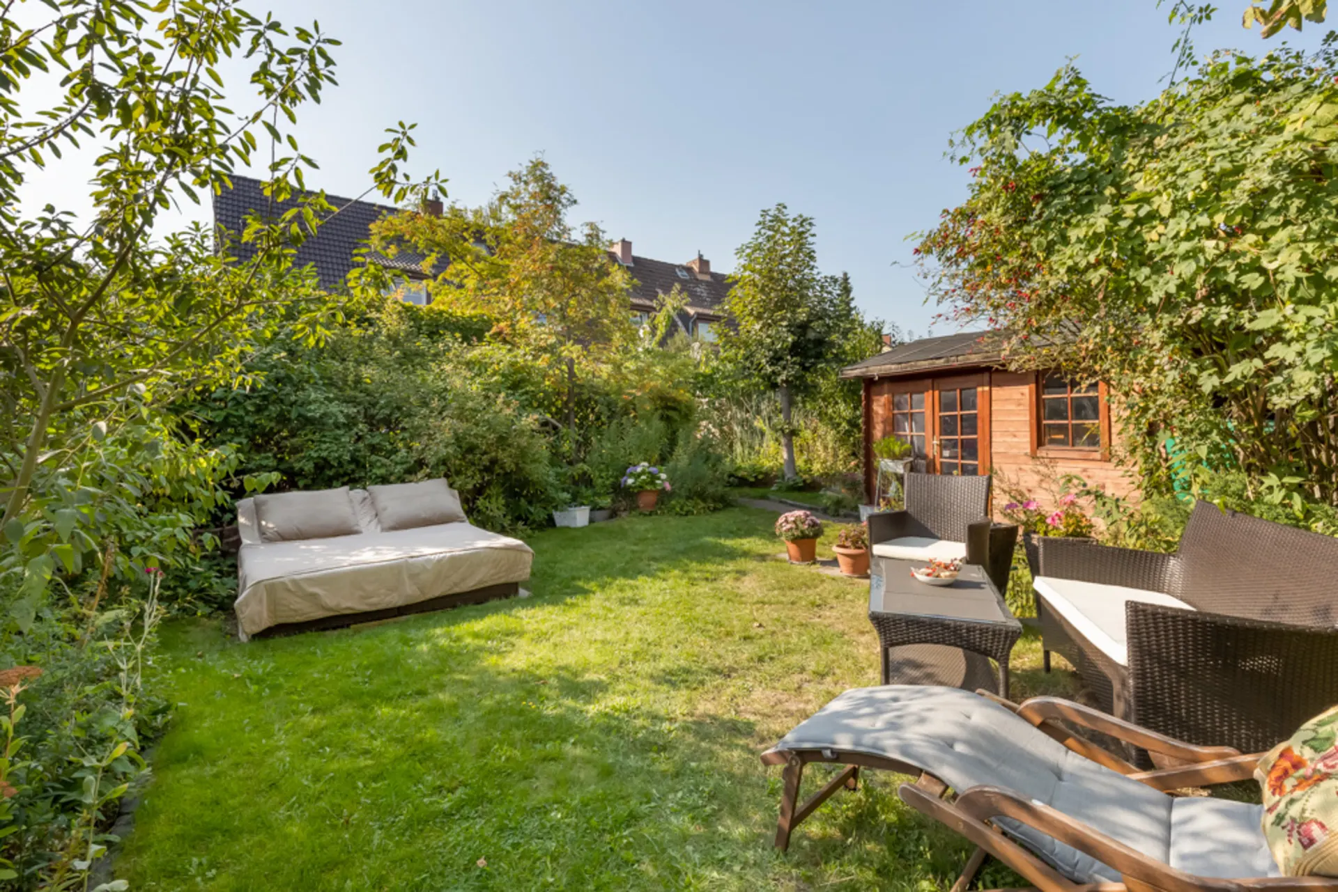 Garten mit Sitzgruppe, Sonnenliege, Sofa und Holzhaus, umgeben von grünen Pflanzen.