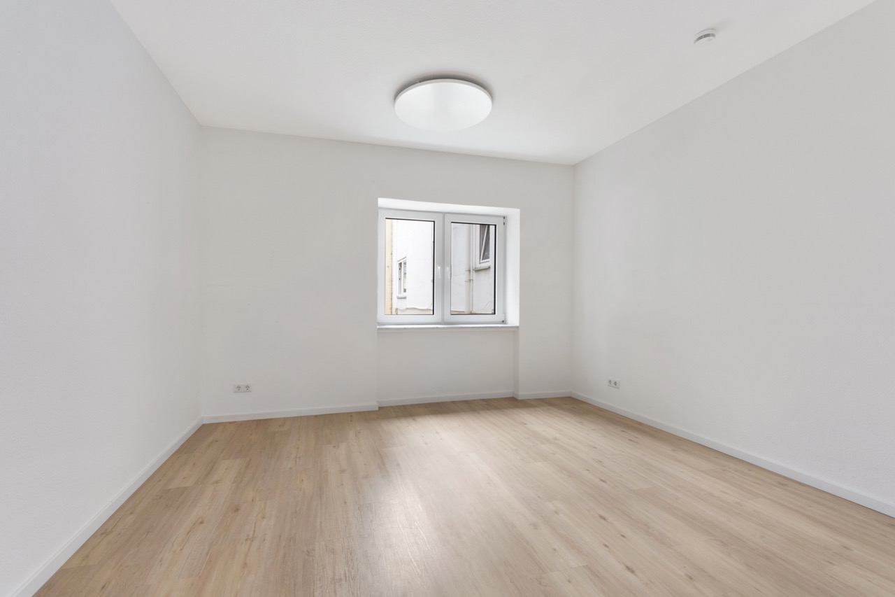 Leerer Raum mit hellem Holzboden, weißen Wänden und einem Fenster in der Mitte.