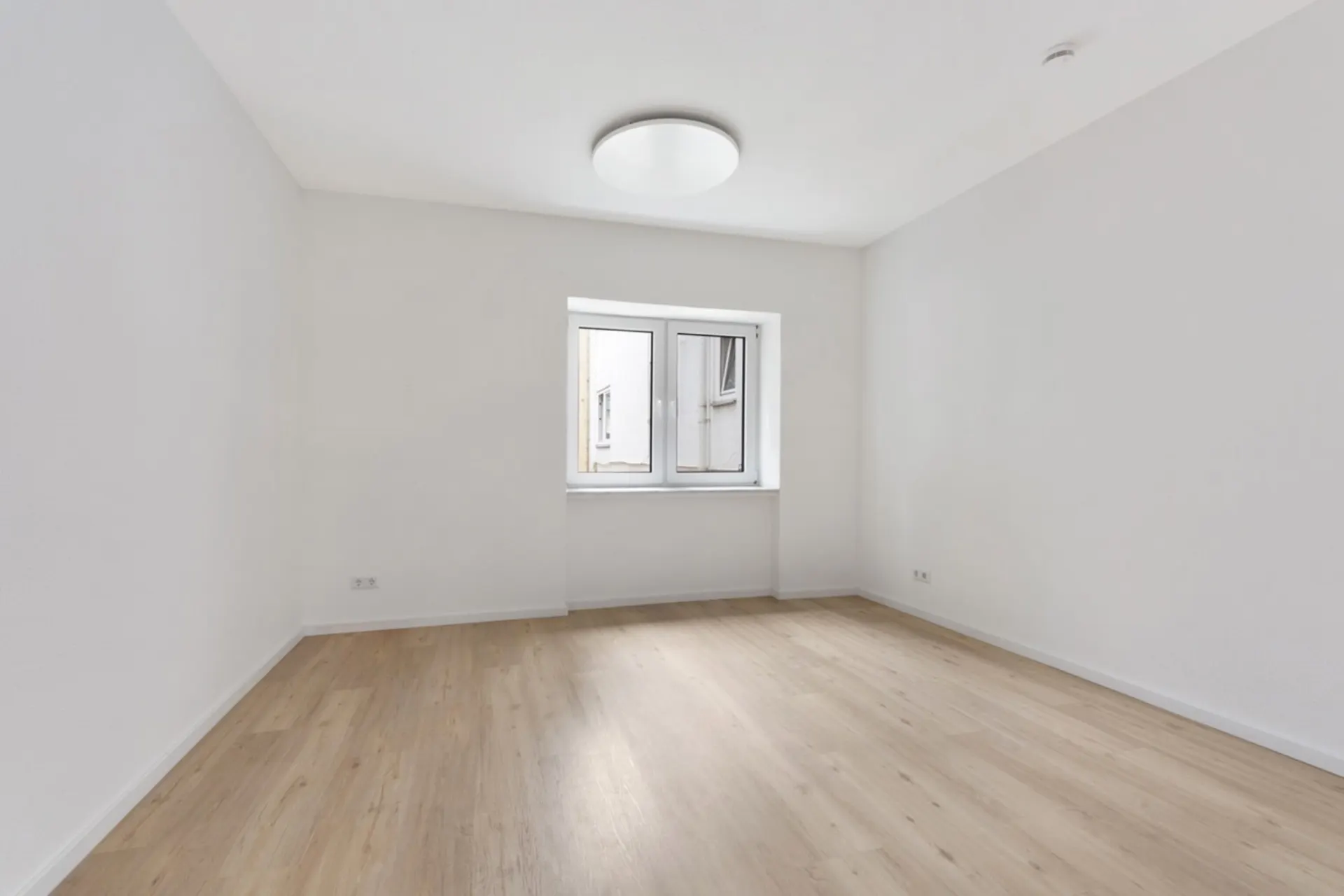 Leerer Raum mit hellem Holzboden, weißen Wänden und einem Fenster in der Mitte.