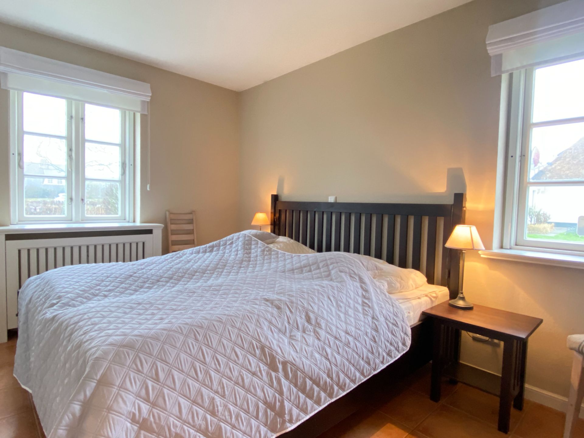 Helles Schlafzimmer mit großem Bett, zwei Nachttischen mit Lampen und zwei Fenstern.
