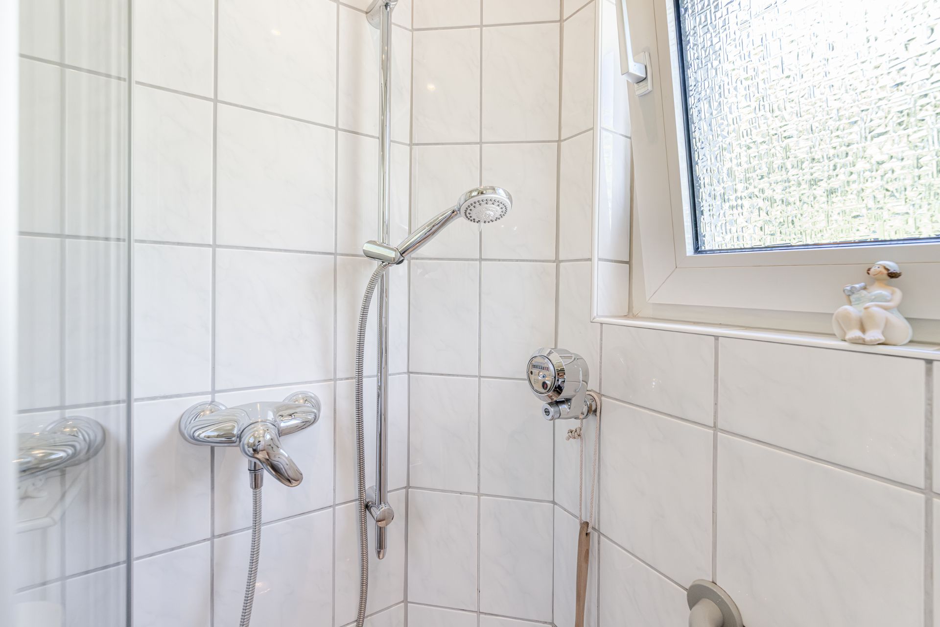 Ecke einer gefliesten Dusche mit Duschkopf, Fenster und Keramikfigur auf der Fensterbank.