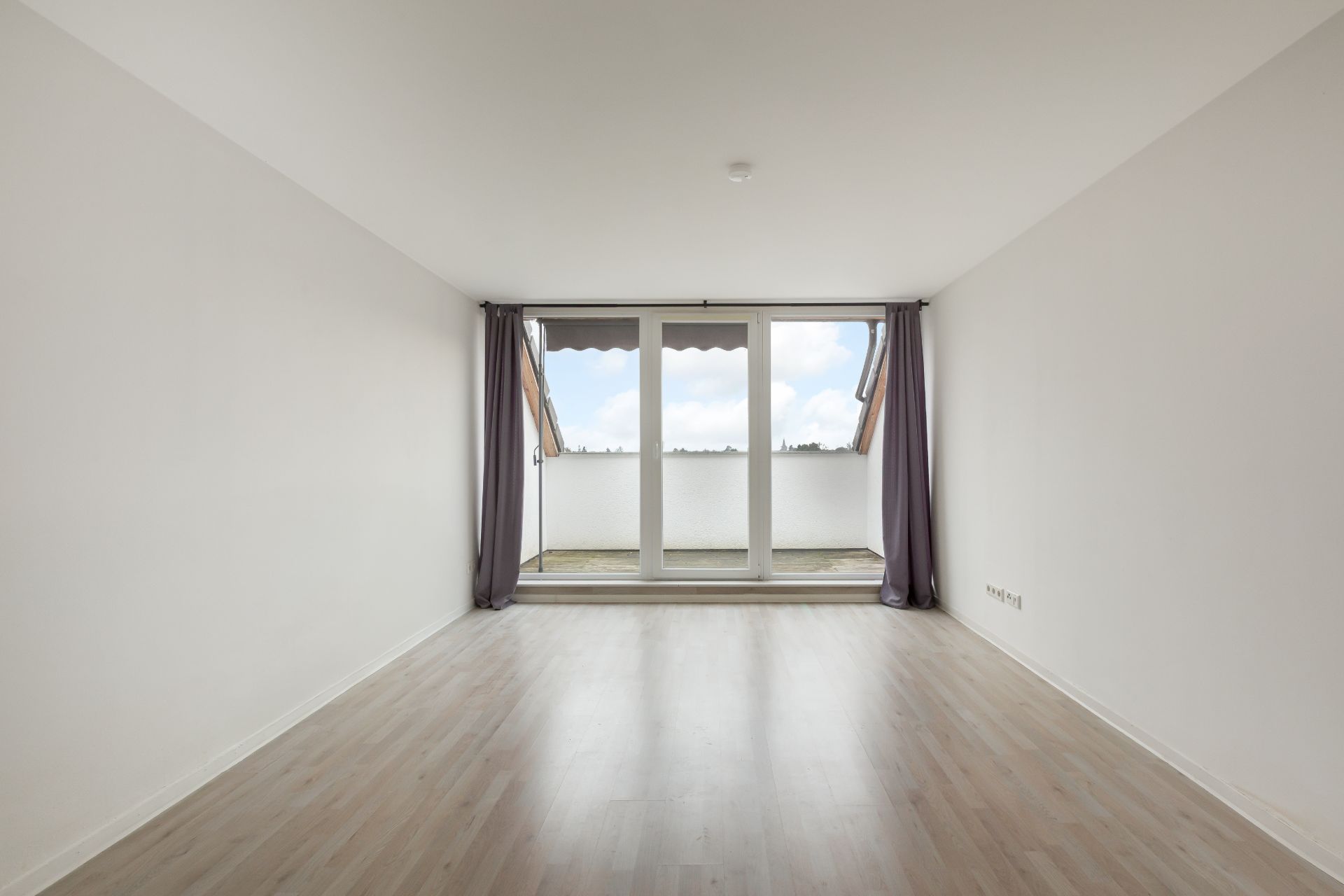 Leeres Zimmer mit hellem Holzboden, bodentiefen Fenstern und Balkonzugang.