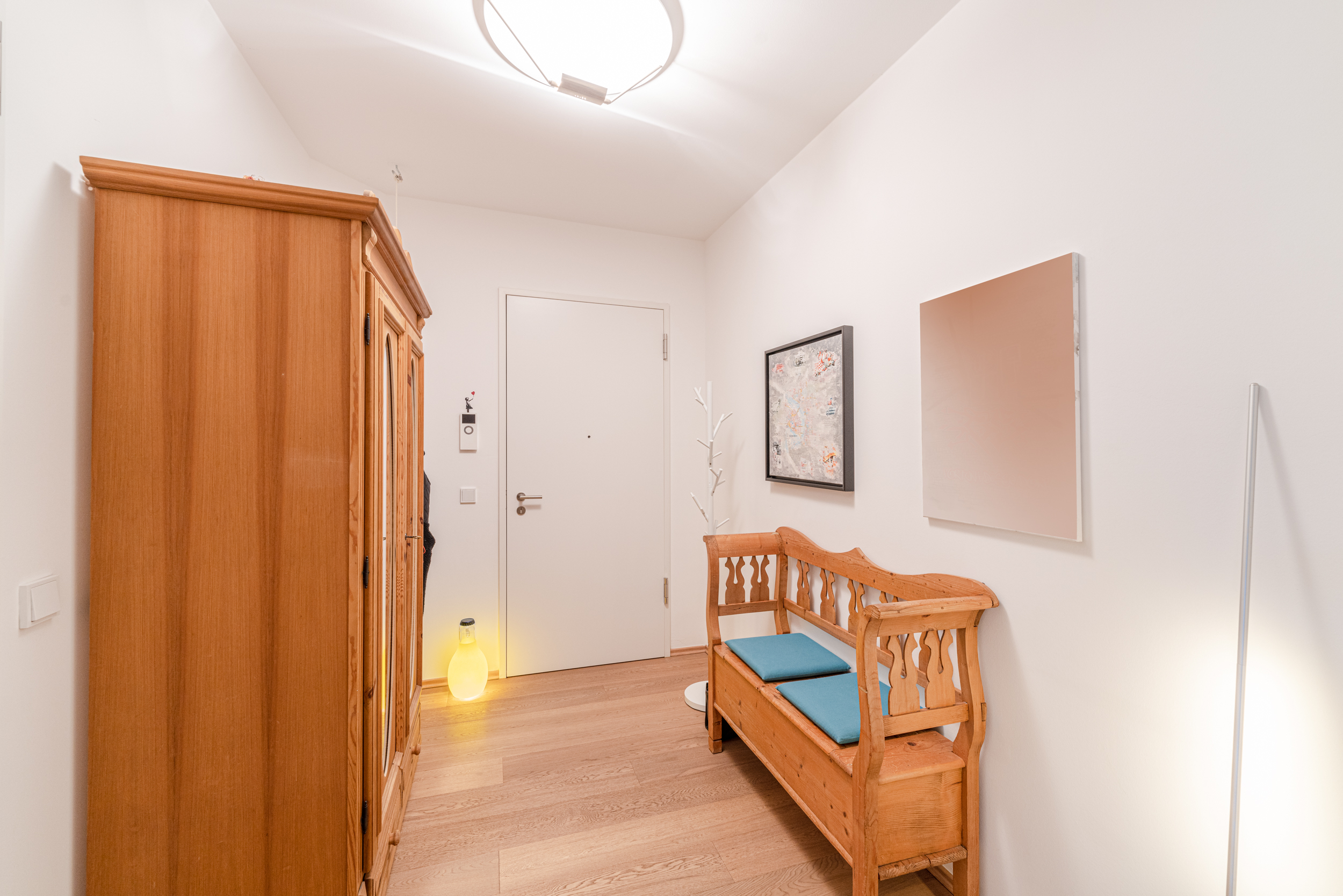 Heller Flur mit Holzbank, großem Schrank, Wandbildern und weißer Eingangstür.
