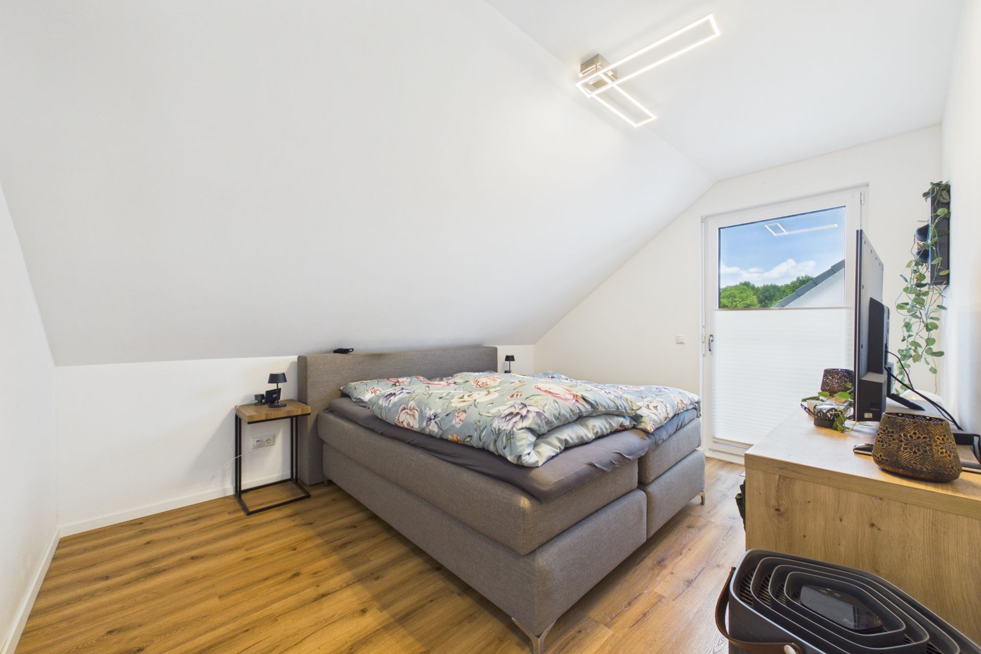 Helles Schlafzimmer mit Doppelbett, Holzboden, Kommode, Fernseher und Zugang zu einem Fenster mit Ausblick.