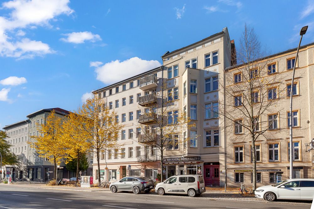 Mehrstöckiges Wohnhaus mit Balkonen, Bäumen vor dem Gebäude und parkenden Autos an der Straße.