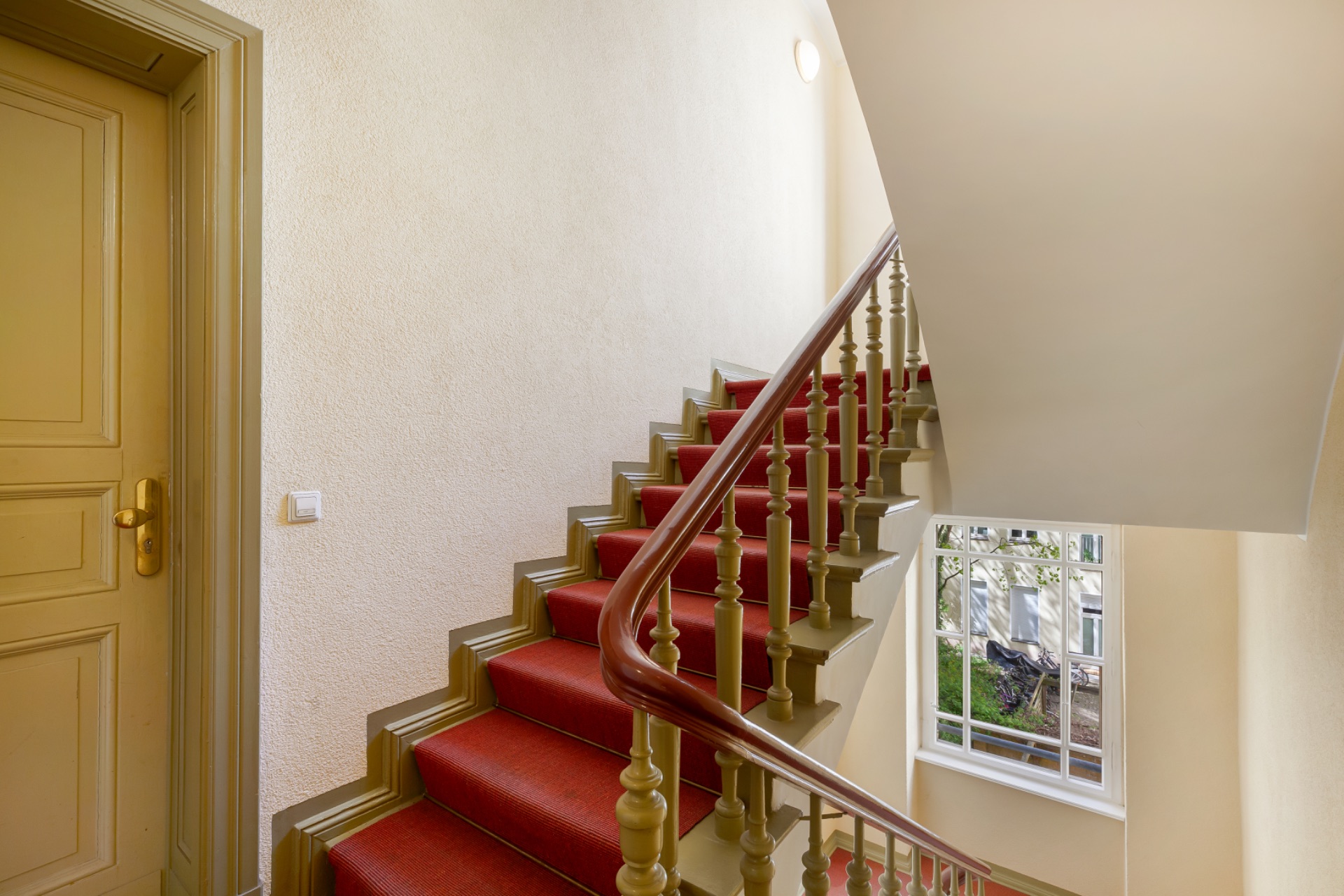 Treppenhaus mit rotem Teppich, Geländer und großem Fenster, daneben eine beige Tür.