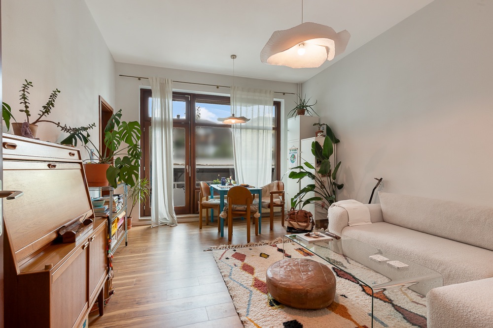 Helles Wohnzimmer mit Sofa, Glastisch, Pflanzen und Esstisch am großen Fenster.