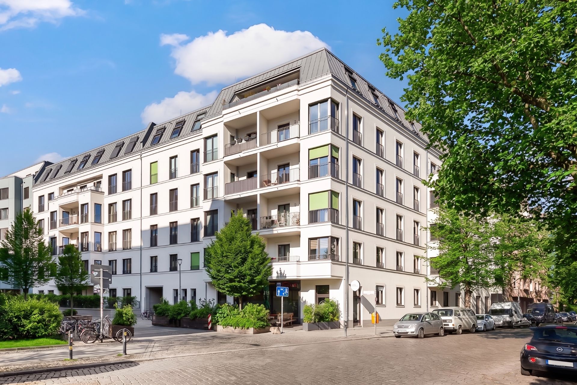 Modernes Mehrfamilienhaus mit Balkonen an einer Straßenecke, umgeben von Bäumen und parkenden Autos.