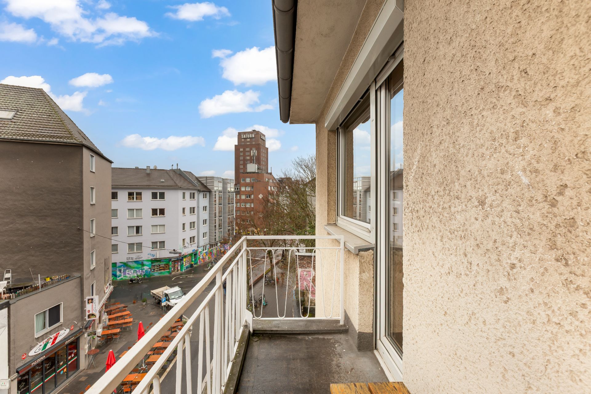 Kleiner Balkon mit weißem Geländer und Ausblick auf eine Straße mit Wohnhäusern.
