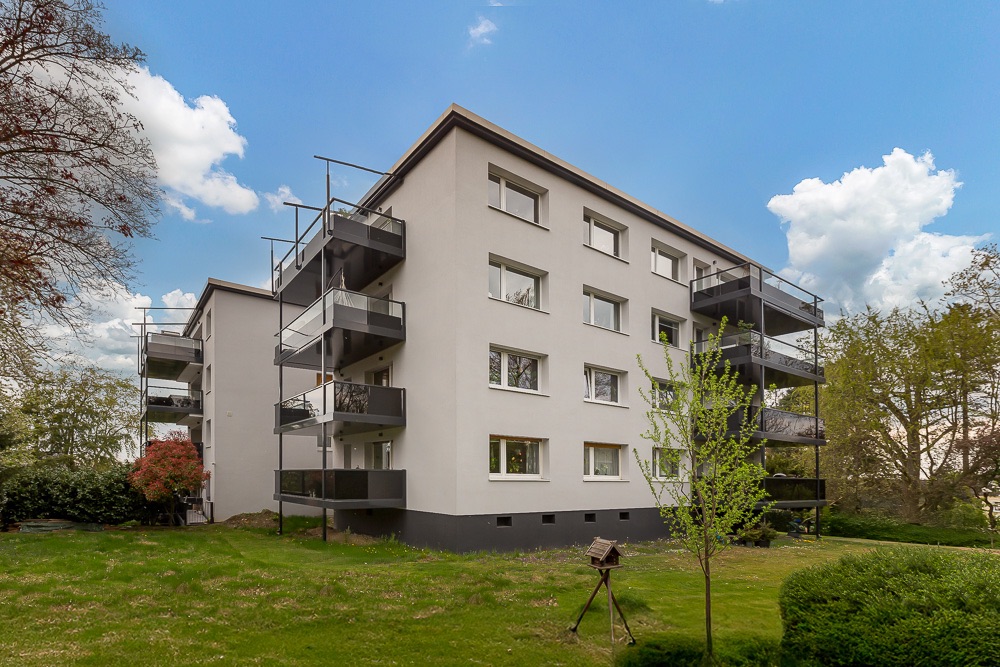 Mehrfamilienhaus mit mehreren Balkonen, umgeben von Rasen und Bäumen an einem sonnigen Tag.