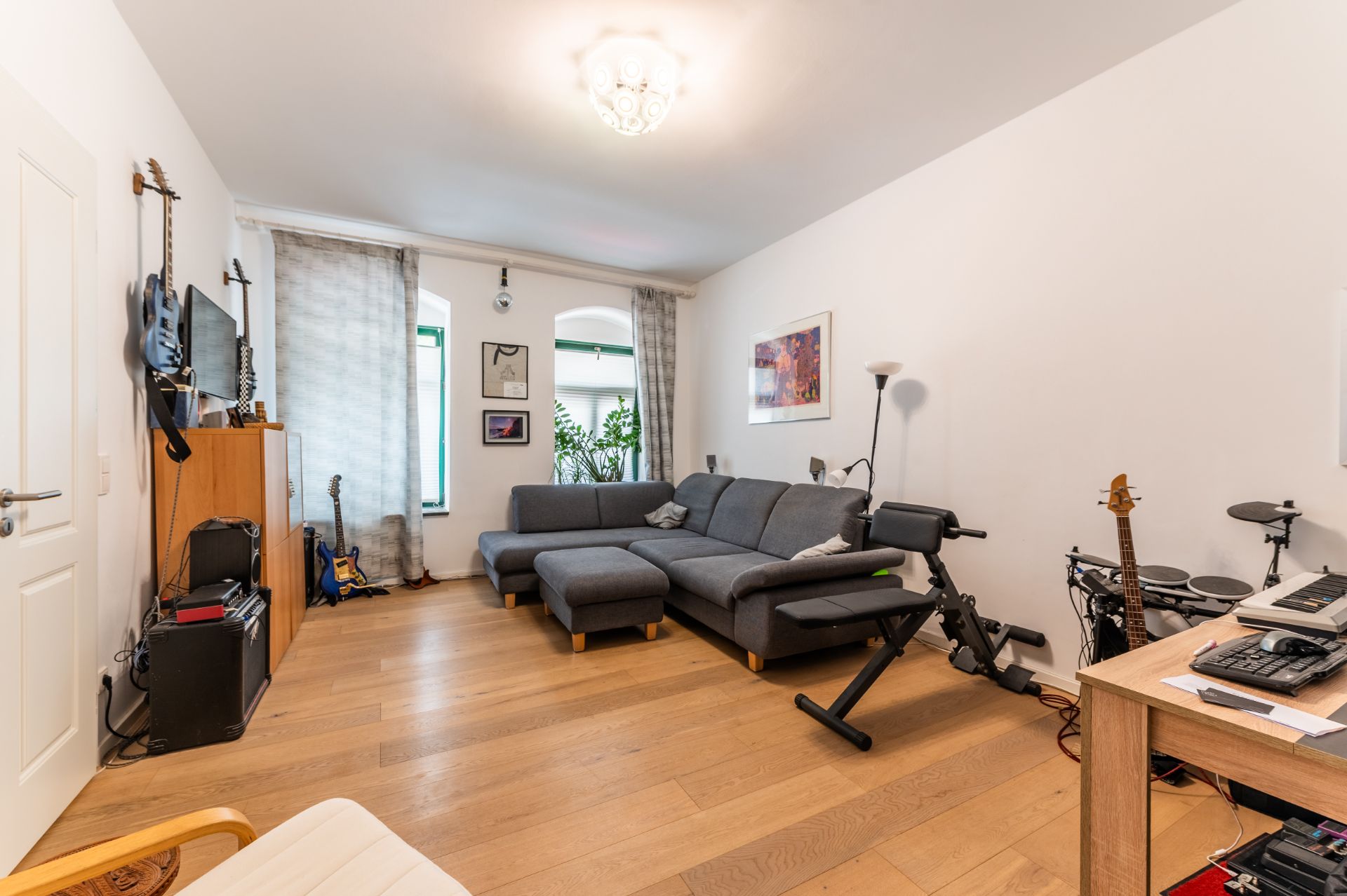Wohnzimmer mit grauem Ecksofa, Musikinstrumenten, Holzboden und Schreibtisch.