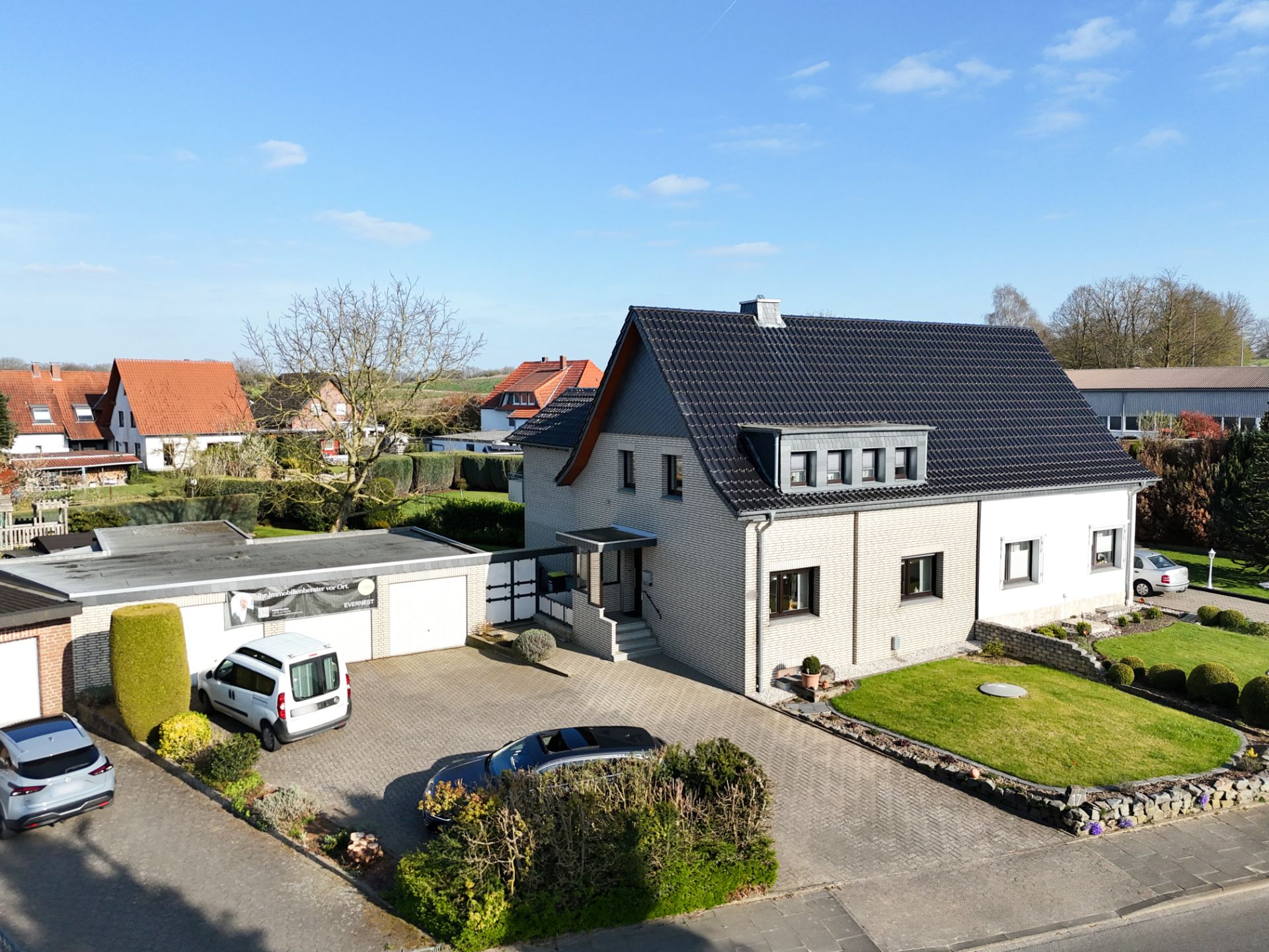 Modernes Einfamilienhaus mit dunklem Dach, gepflegtem Garten und mehreren geparkten Autos in der Einfahrt.