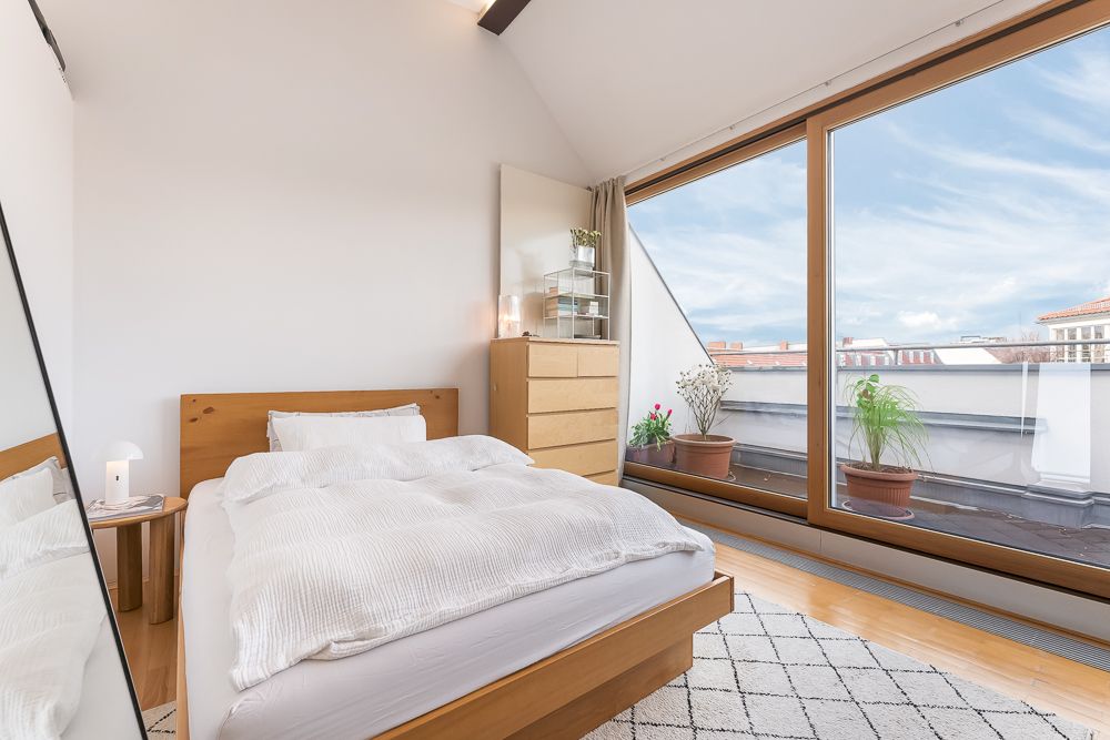 Helles Schlafzimmer mit Holzbett, Kommode, großem Fenster und Balkon mit Pflanzen.