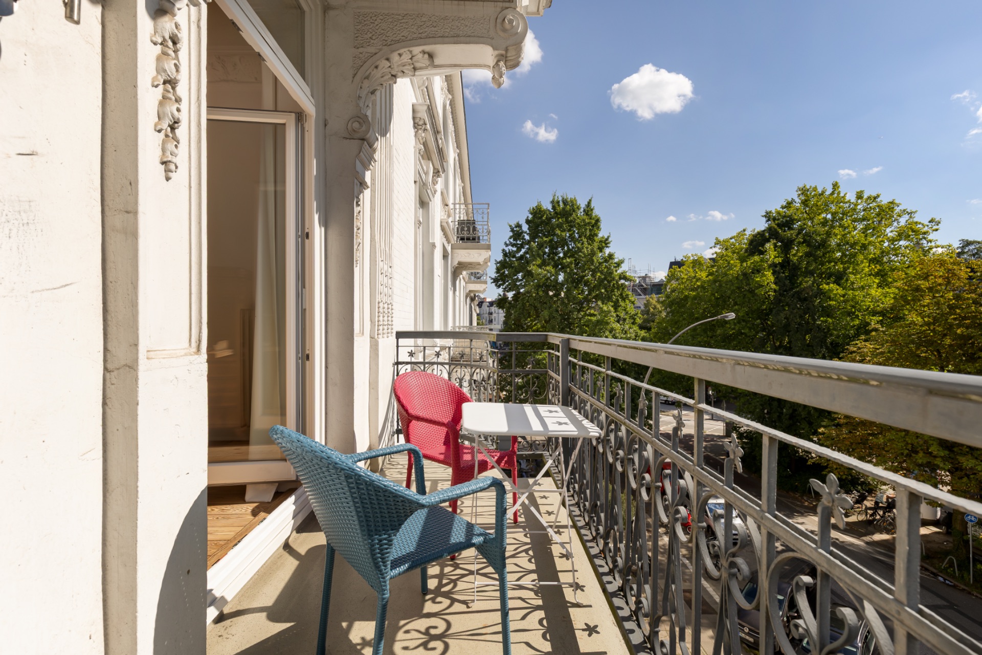 Balkon mit zwei bunten Stühlen und einem kleinen Tisch, Blick auf Bäume und Straße.