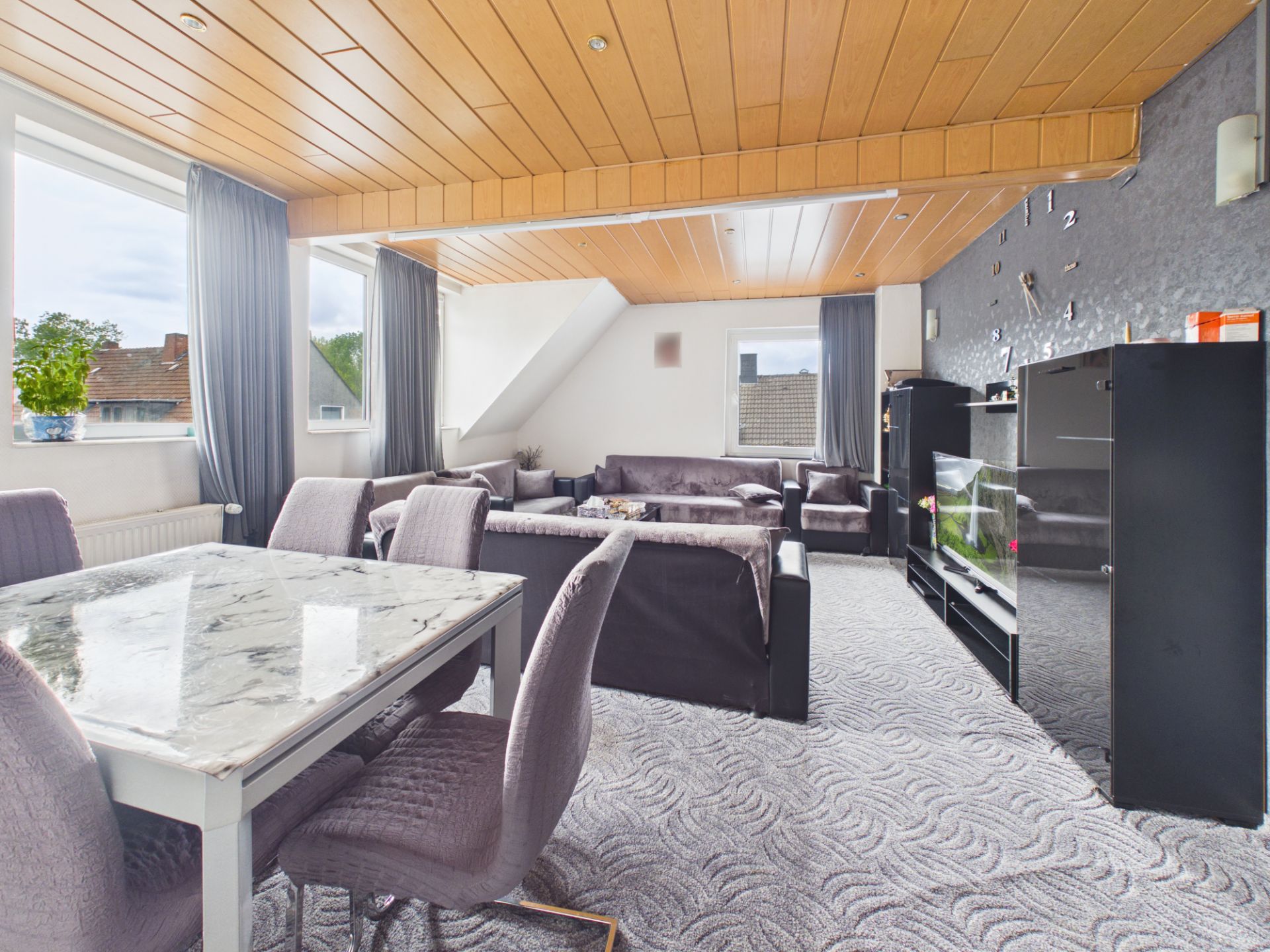 Wohnzimmer mit mehreren Sofas, Esstisch und großem Fenster, Holzdecke und grauen Vorhängen.