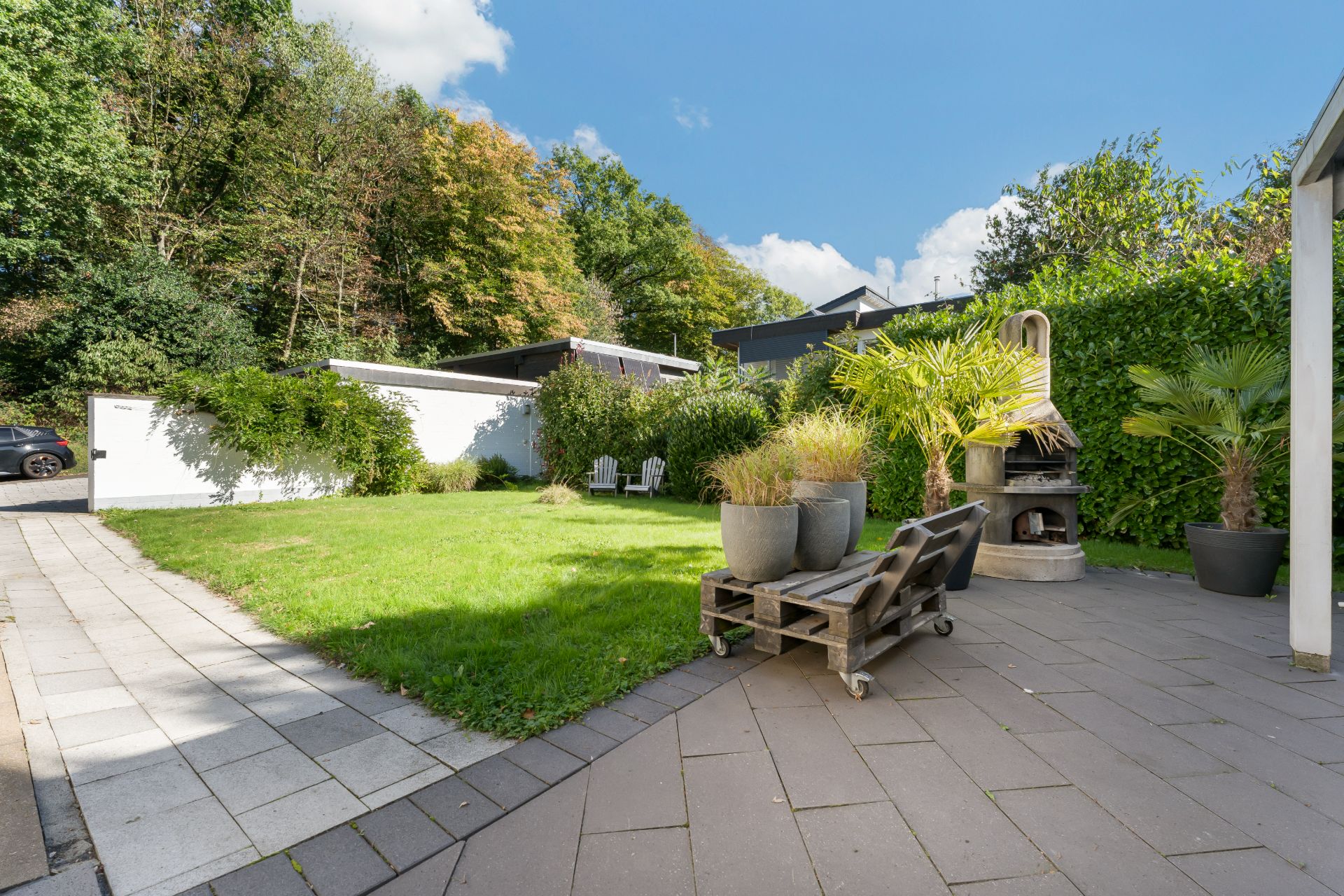 Garten mit Rasen, Terrasse, Sonnenliegen, Pflanzenkübeln und festem Grillplatz.
