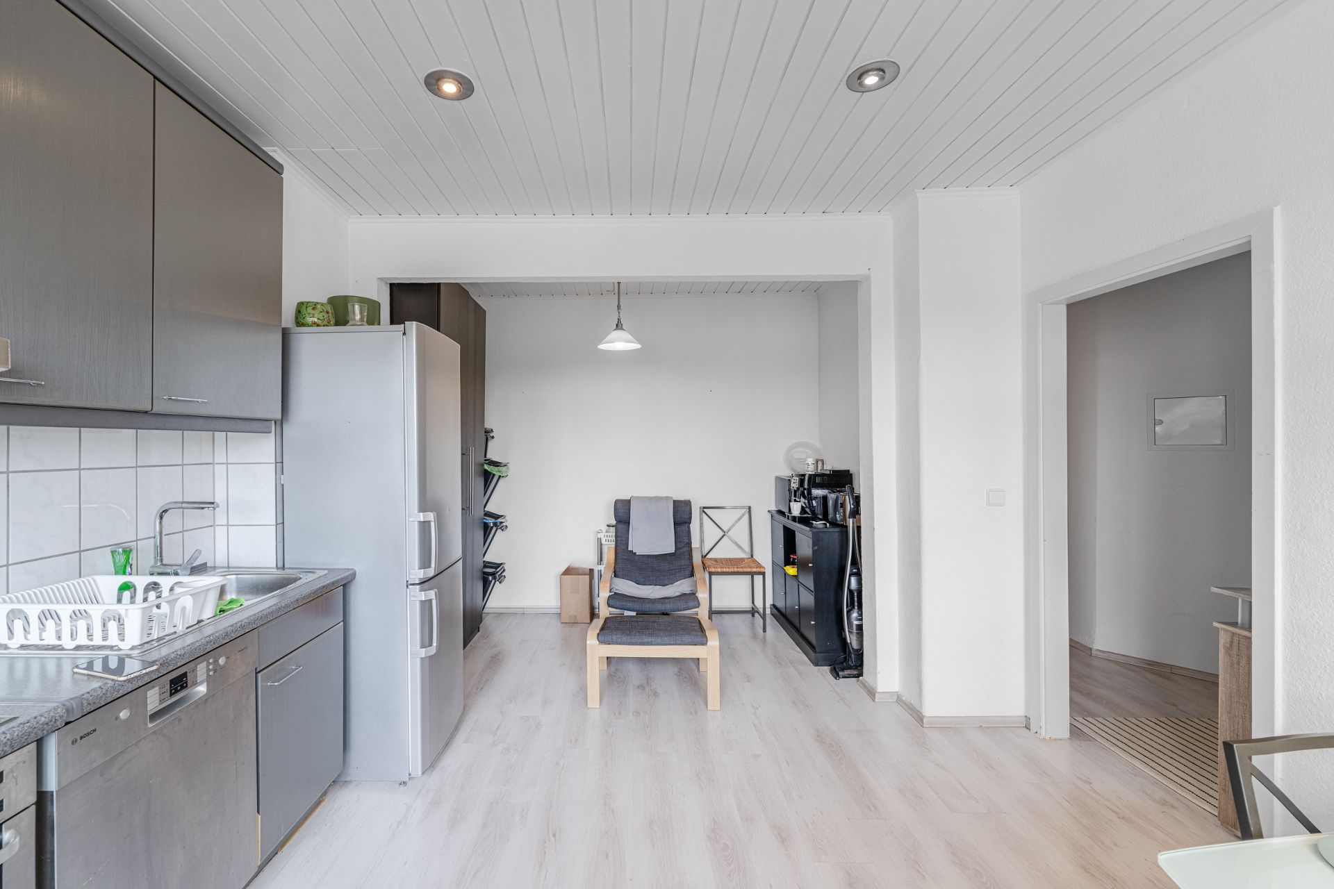 Helle Küche mit Spüle, Kühlschrank, Sessel und kleinem Regal unter Hängelampe.