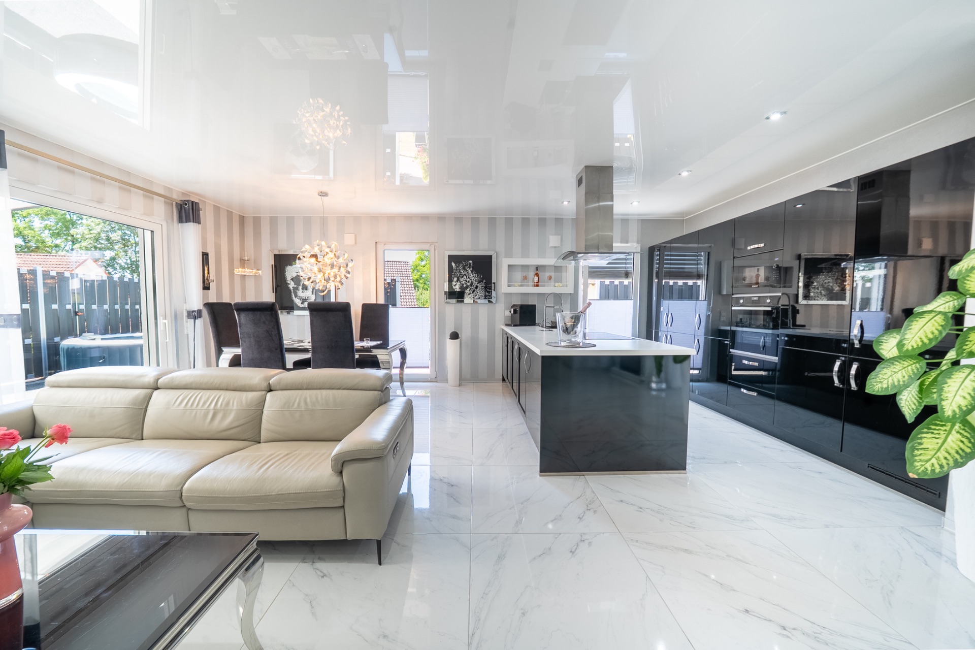 Modernes Wohnzimmer mit heller Ledercouch, Essbereich und offener, schwarzer Küche auf Marmorboden.