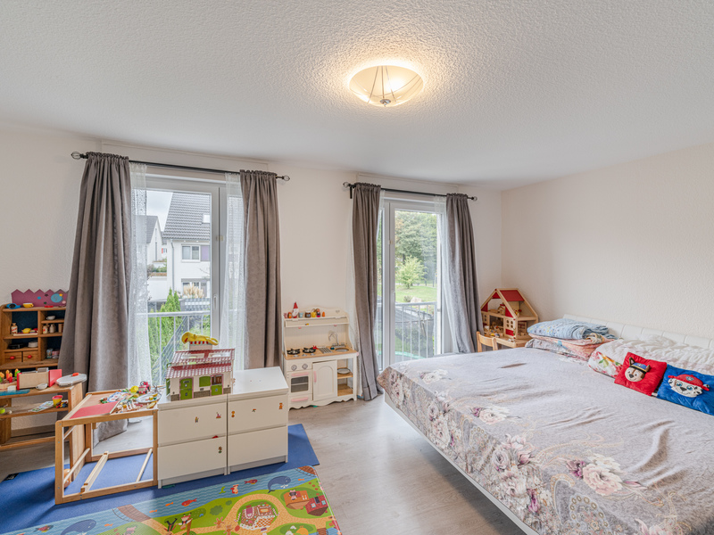 Helles Kinderzimmer mit Spielzeug, großem Bett, Kindermöbeln und zwei Fenstern mit grauen Vorhängen.