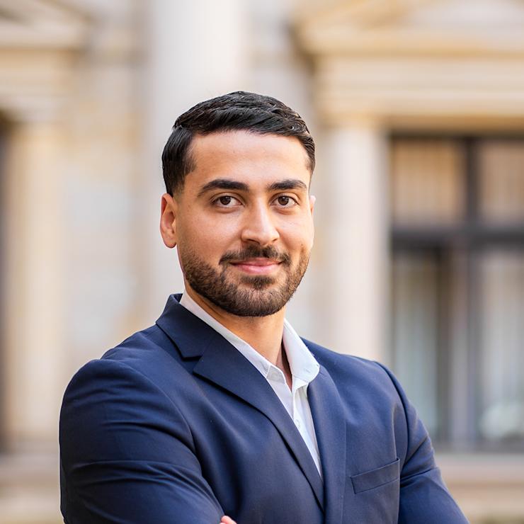 Ein Mann im blauen Anzug steht mit verschränkten Armen vor einem Gebäude.