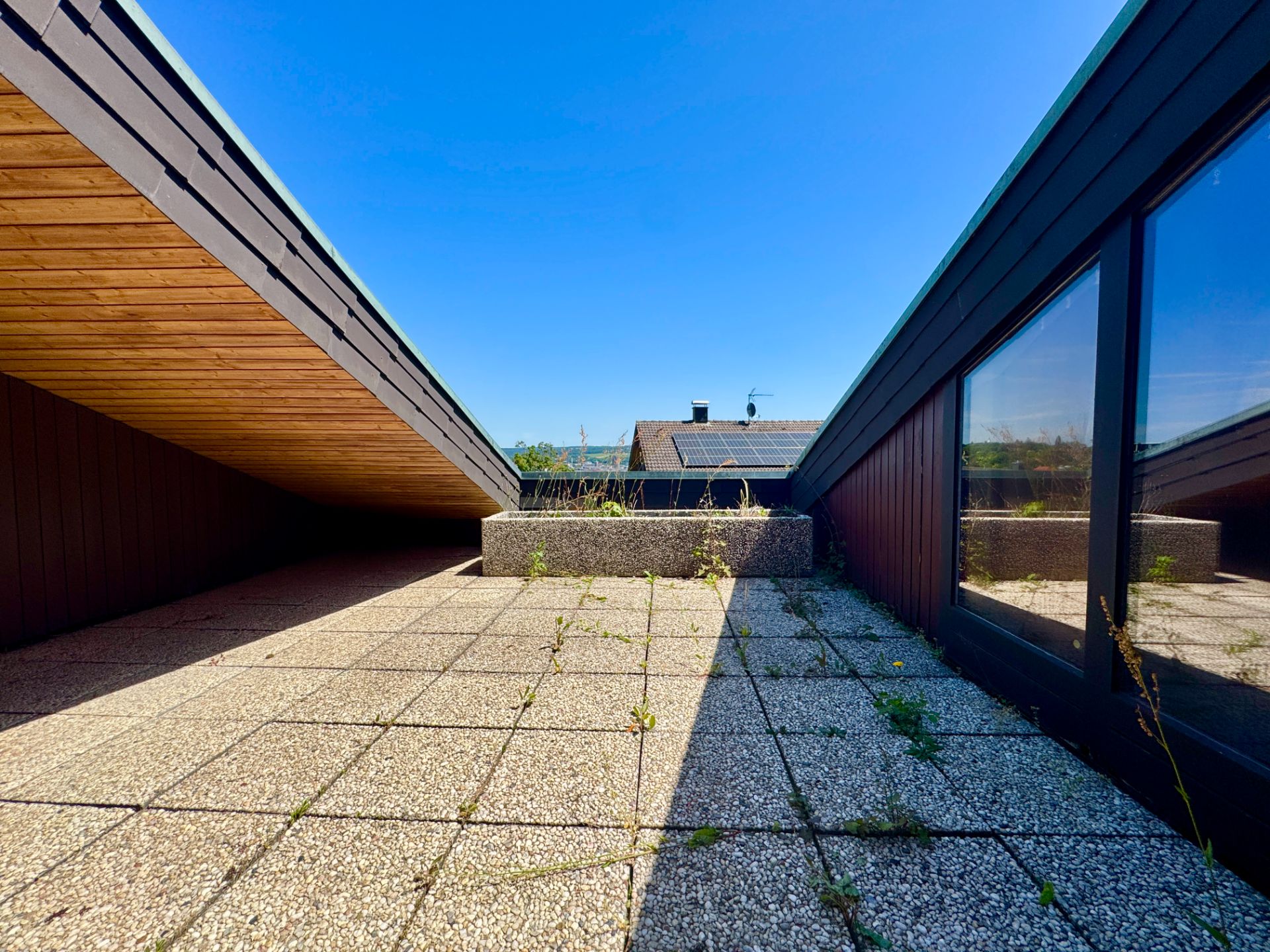 Dachterrasse mit Steinfliesen, niedrigem Geländer und großen Fenstern, blauer Himmel im Hintergrund.