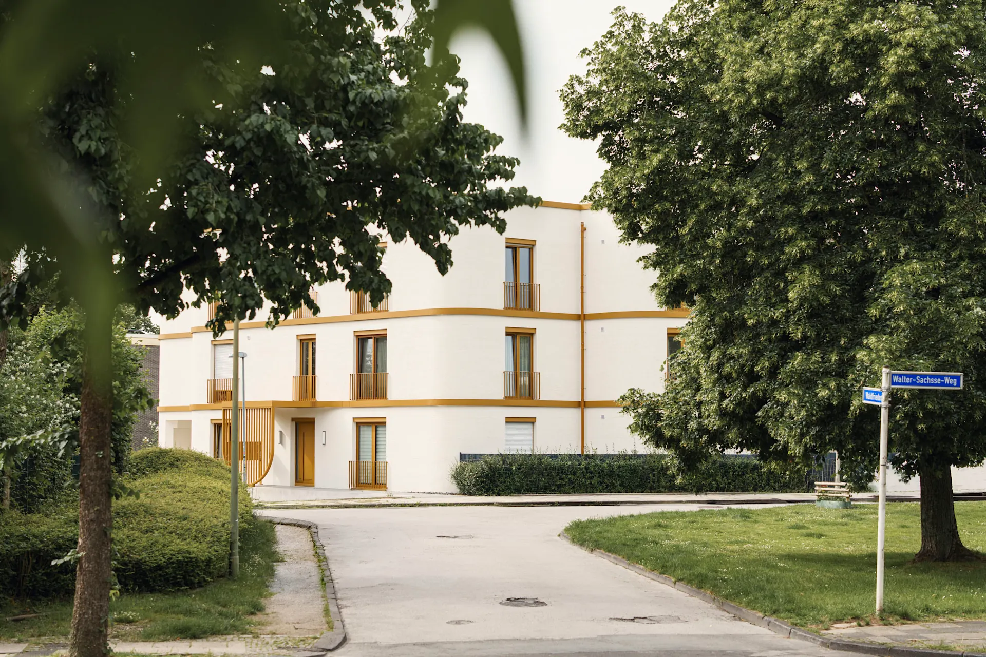 Modernes, dreistöckiges Wohnhaus mit weißer Fassade an einer ruhigen Straße, umgeben von Bäumen.