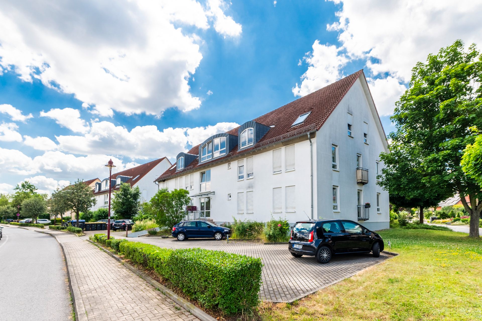 Wohngebäude mit mehreren Autos auf Parkplatz und Bäumen im Vordergrund bei blauem Himmel.