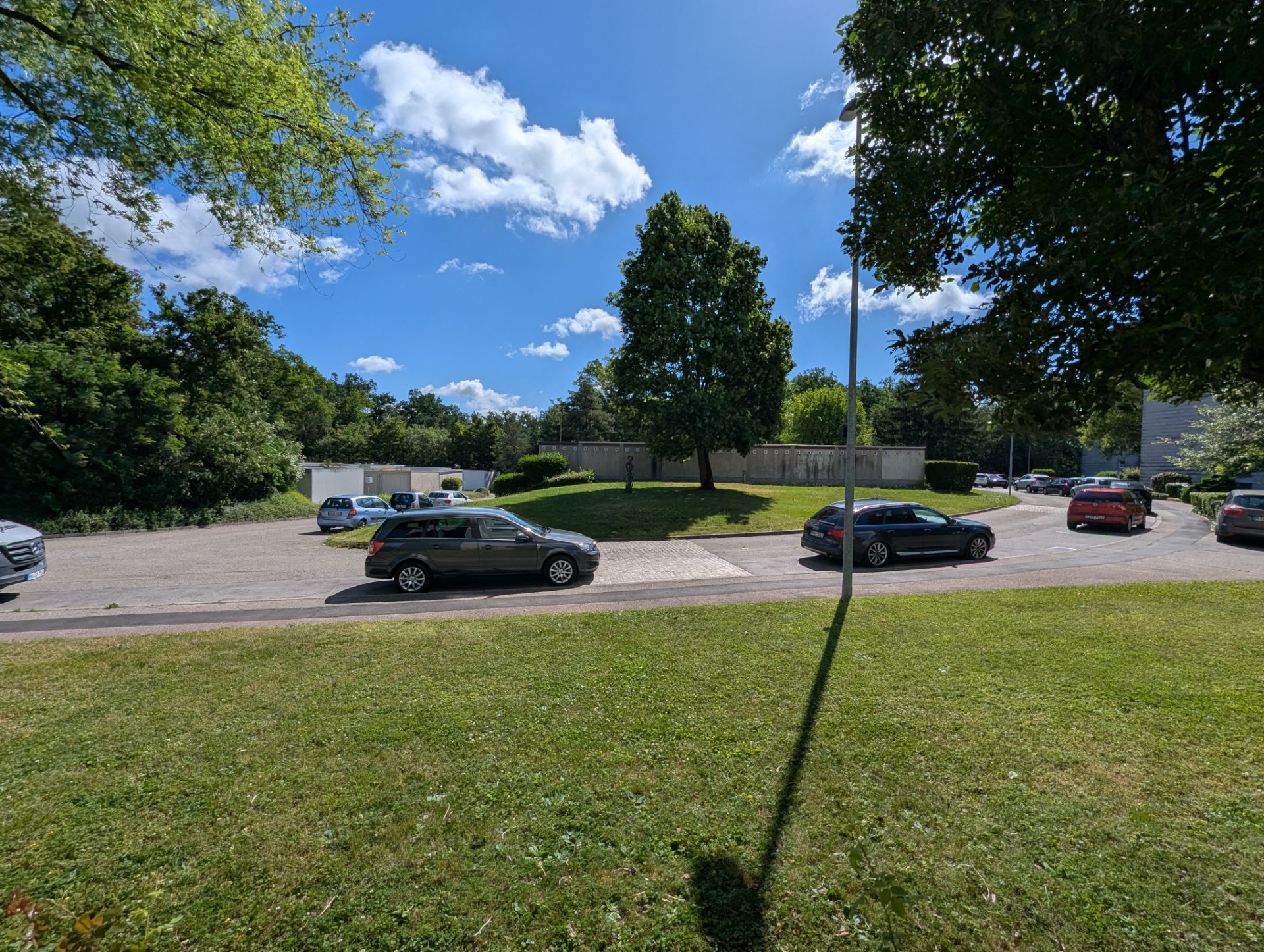 Parkplatz mit mehreren Autos, Bäumen und einer Rasenfläche bei sonnigem Wetter.