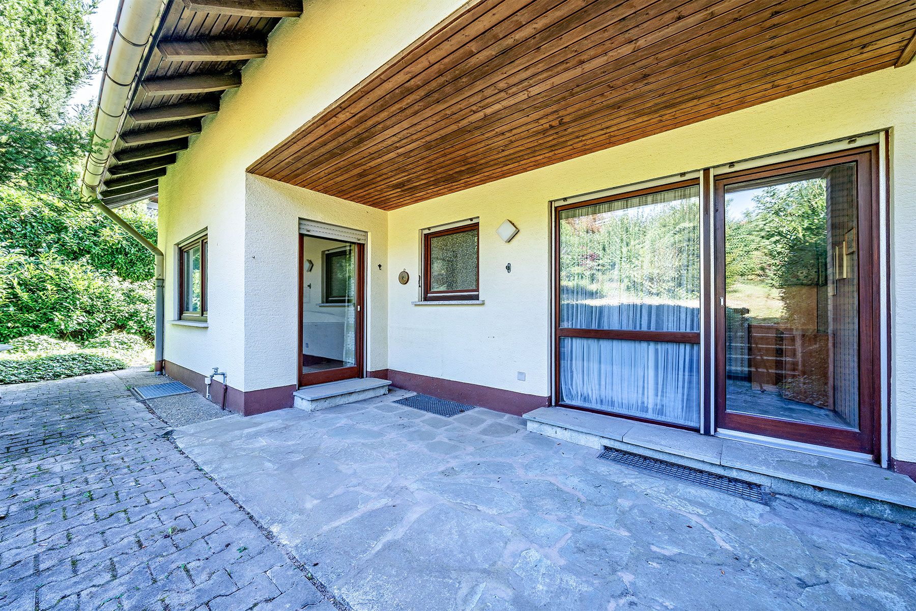 Überdachte Terrasse mit Steinboden, Fenstern und zwei Glastüren eines Wohnhauses.