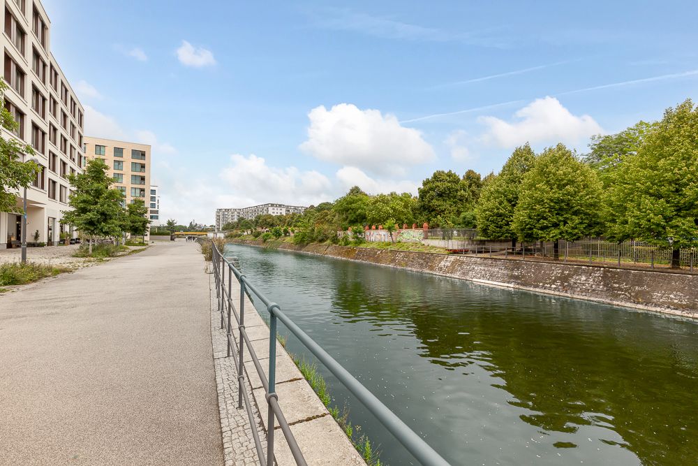 Uferweg mit Geländer neben einem Fluss, links moderne Wohngebäude, rechts grüne Bäume.