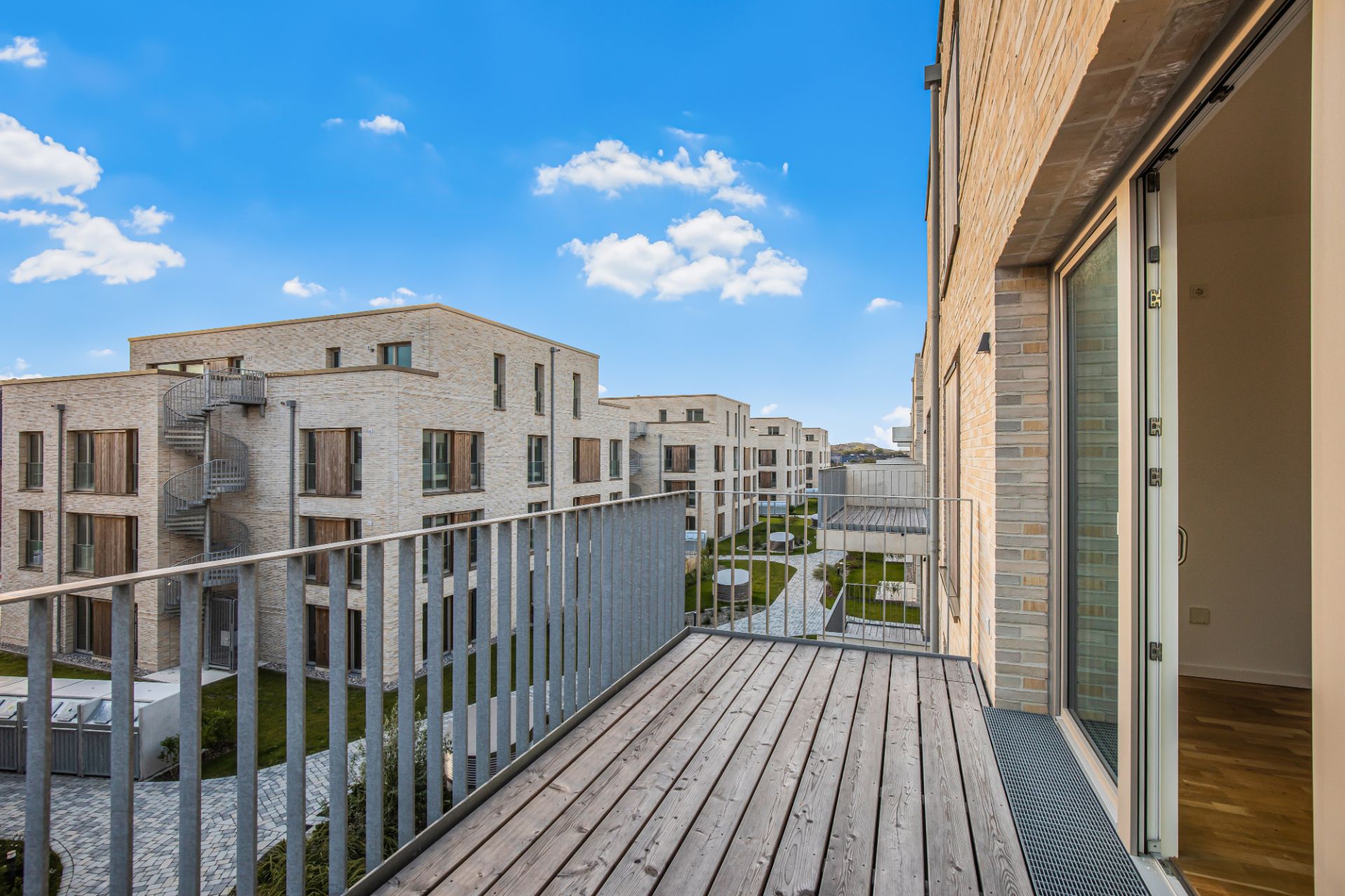 Blick von einem Balkon auf moderne Wohnhäuser mit Terrasse und Garten, blauer Himmel im Hintergrund.