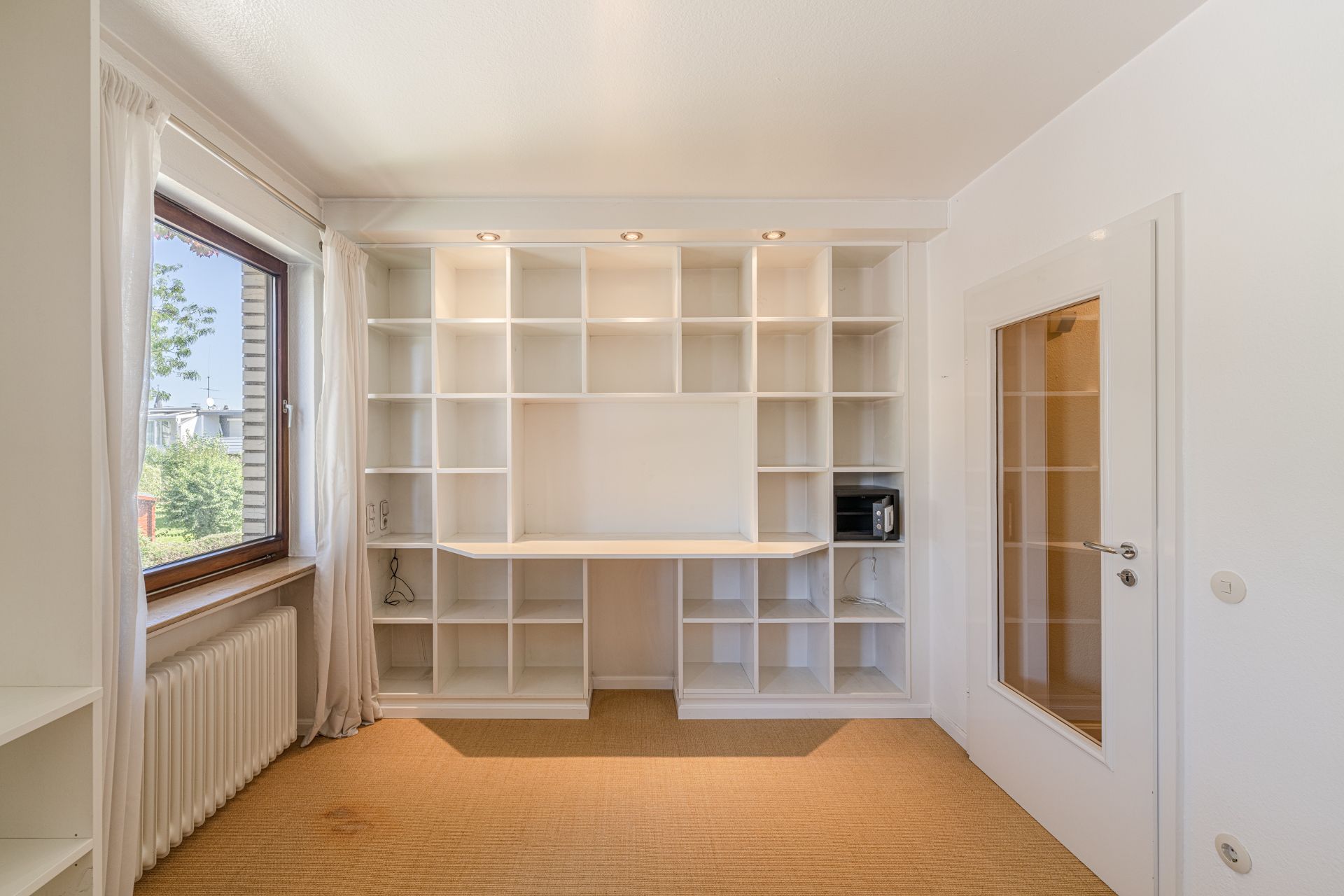 Helles Zimmer mit großem weißen Regal, Fenster mit weißen Vorhängen und Glastür.