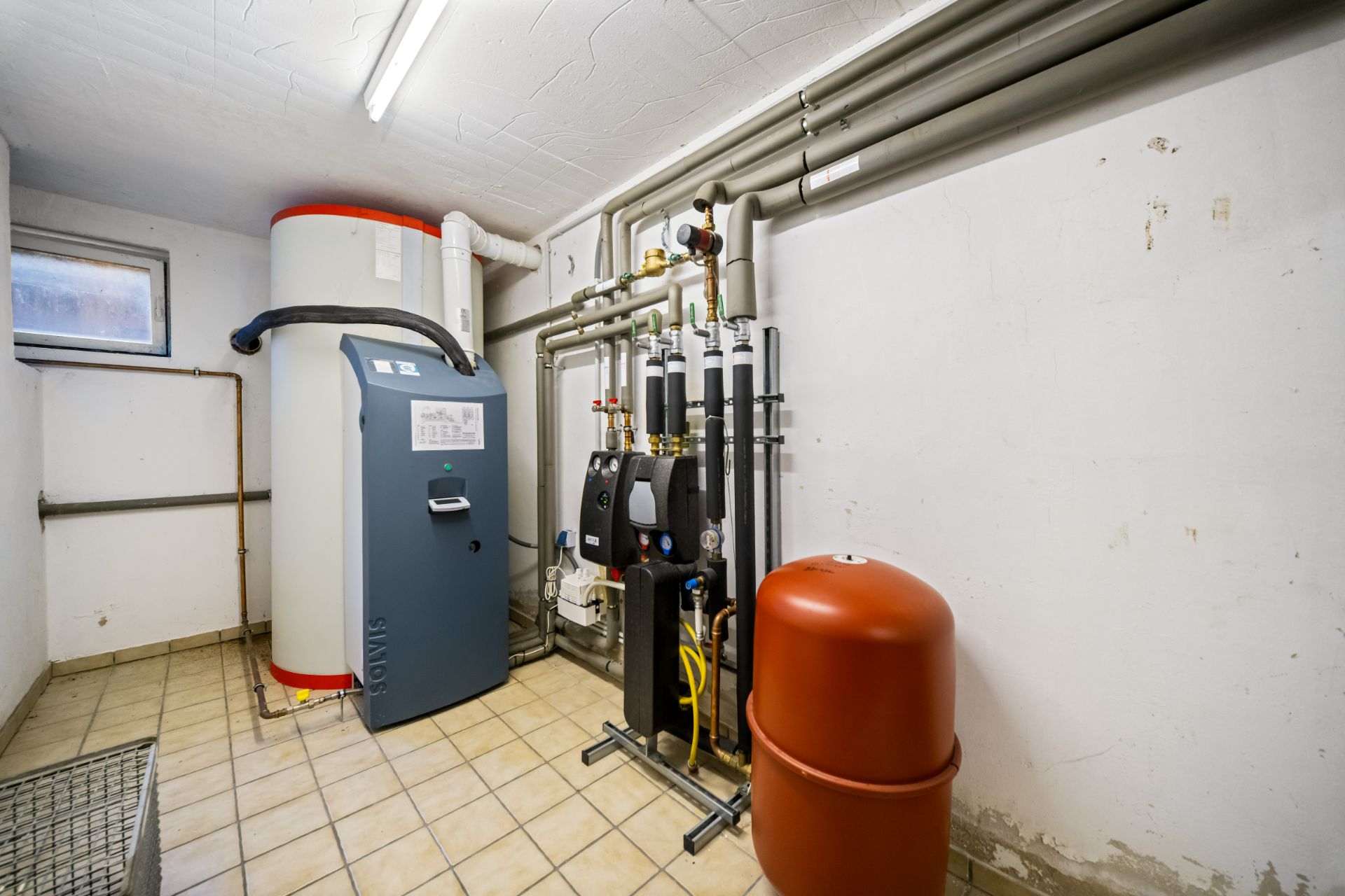 Heizungsraum mit Heizkessel, Speicher und mehreren Rohren an der Wand.