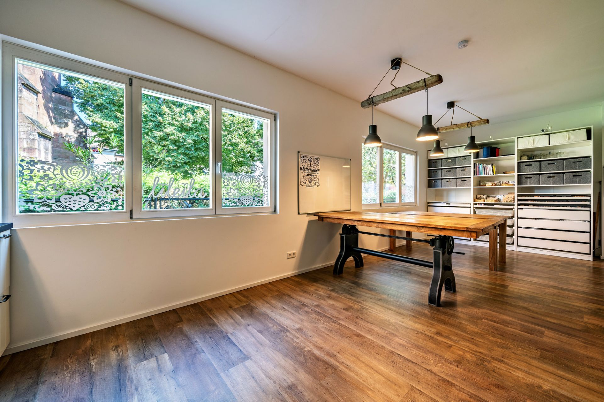 Helles Zimmer mit Holzboden, großem Tisch, Regalen und Fenstern mit Blick ins Grüne.