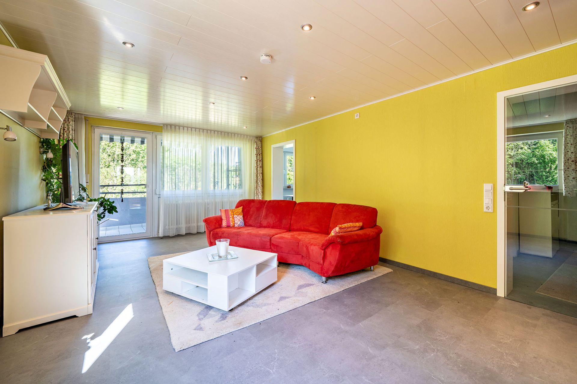 Wohnzimmer mit rotem Sofa, gelber Wand, weißem Tisch und Zugang zum Balkon.
