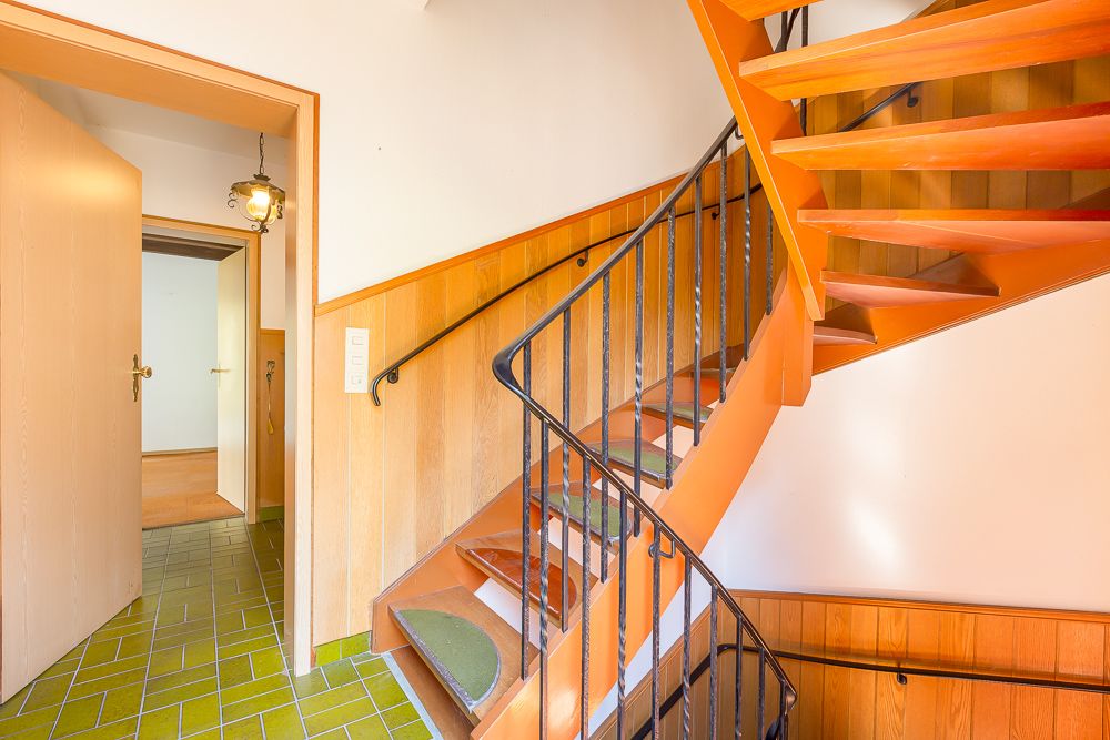 Treppenhaus mit orangener Holztreppe, grünem Fliesenboden und offenen Türen.