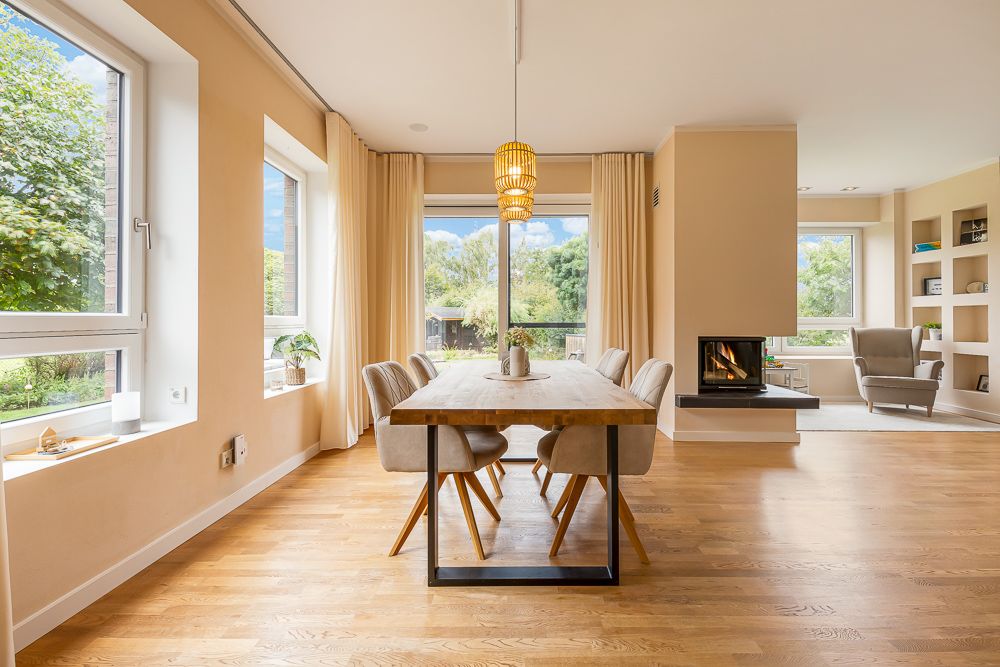 Essbereich mit Holztisch und sechs Stühlen, große Fenster, Kamin und heller Wohnraum sichtbar.
